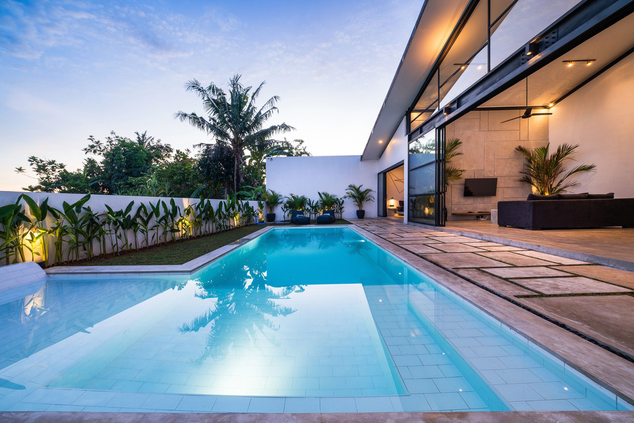 A modern house with a large outdoor pool, surrounded by plants and featuring large windows and a seating area. The sky is clear, indicating it is either early morning or late afternoon.